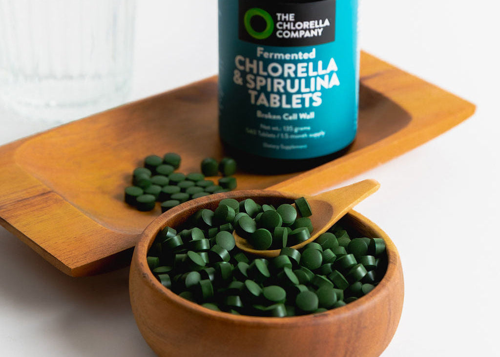 Bottle of Chlorella & Spirulina Tablets on a wooden tray with a bowl of green-blue tablets.