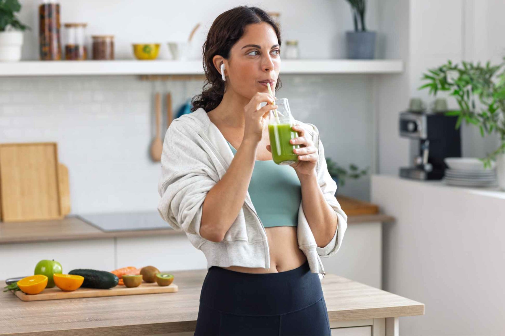 Woman drinking a green smoothie in a kitchen with fruits and a coffee maker in the background
