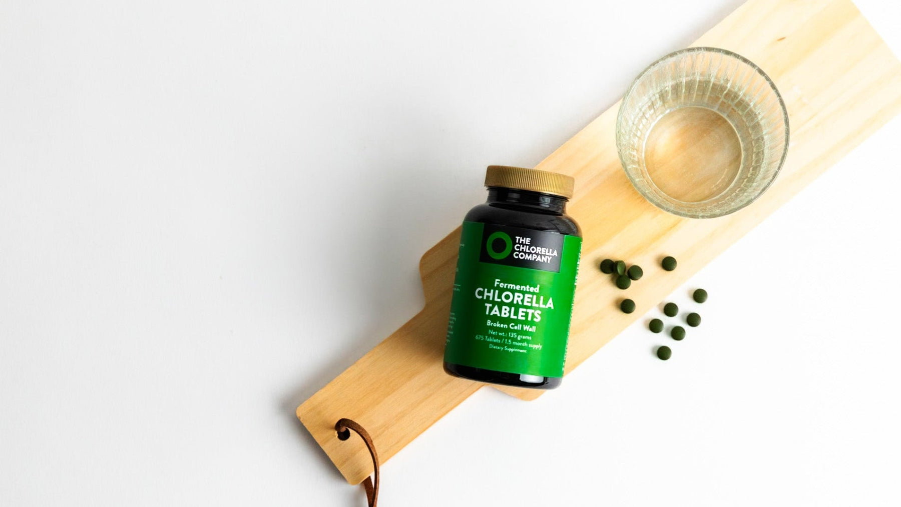 Bottle of chlorella tablets from The Chlorella Company on a wooden board with a glass of water.